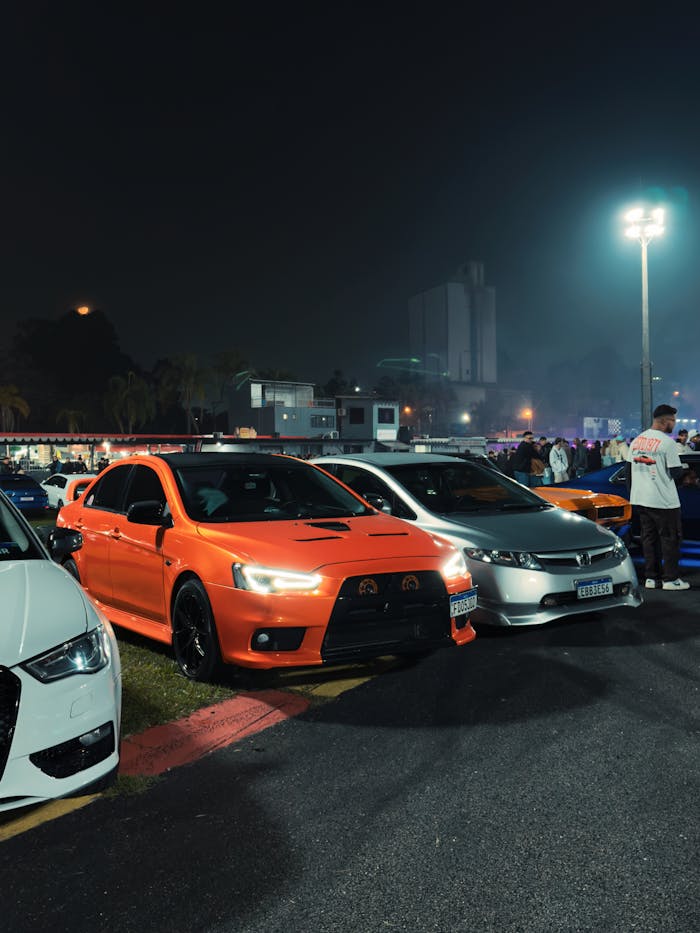 Vibrant night scene of a car meet featuring a bright orange sports sedan, surrounded by various cars.